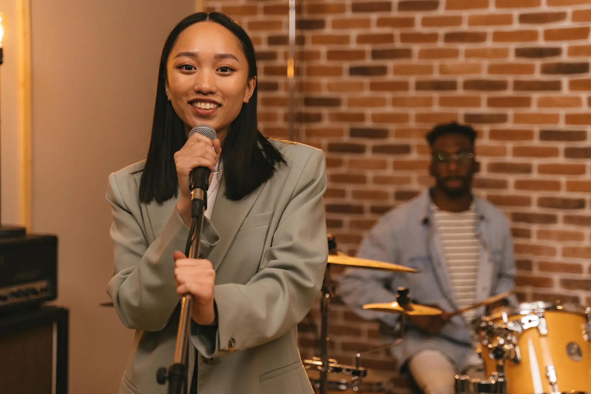 Asian woman singing into a microphone with a drummer in the background, showcasing musical talent.